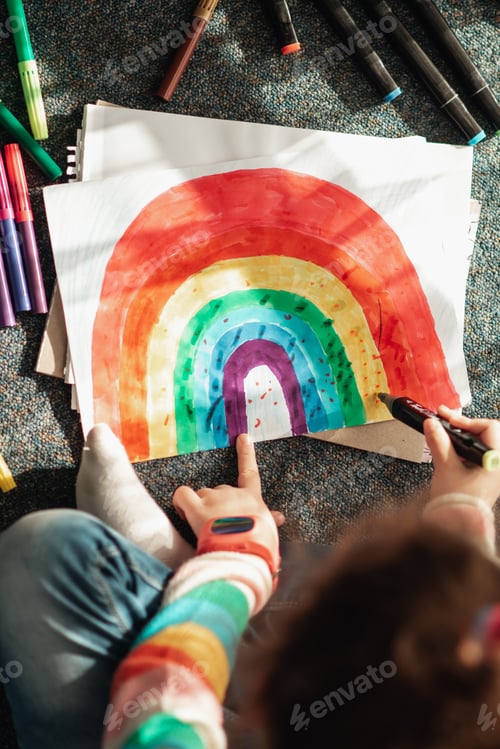 Preview: A little girl sits on the floor in the sun and draws a rainbow with markers.