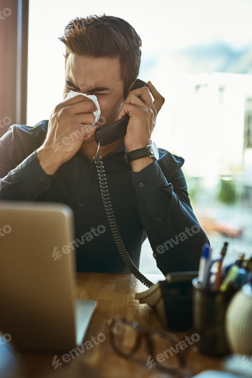 Preview: Shot of a young businessman blowing his nose while speaking on a phone in an office