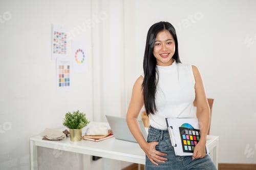 Preview: A smiling asian woman is carrying the clipboard in one hand while posing in front of her work desk.