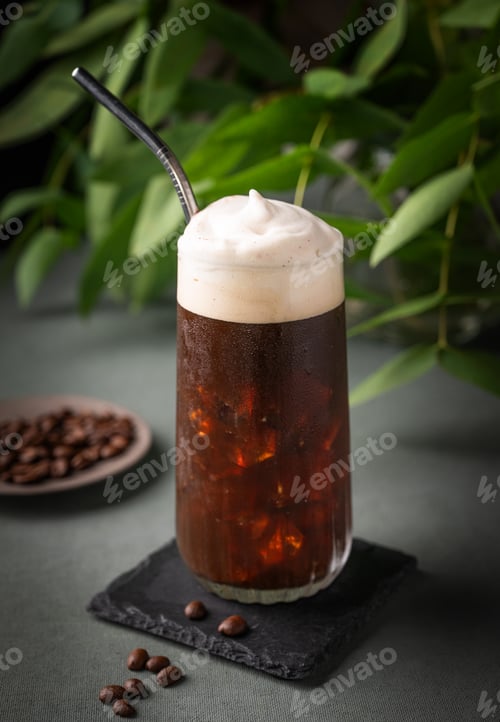 Preview: Iced coffee brew with foam in a glass on a black board on a dark green background with coffee beans