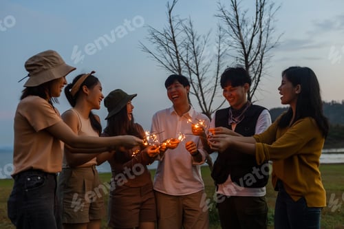 Preview: Happy friends holding fire sparkles. Group of young Travel join in fun at camping party in nature.