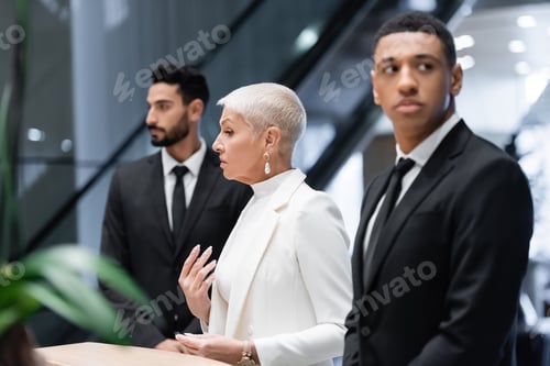 Preview: stylish senior business lady near hotel reception and private multiethnic bodyguards
