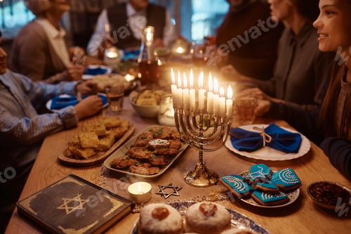 Preview: Lit candles in menorah on dining table with extended Jewish family in the background.