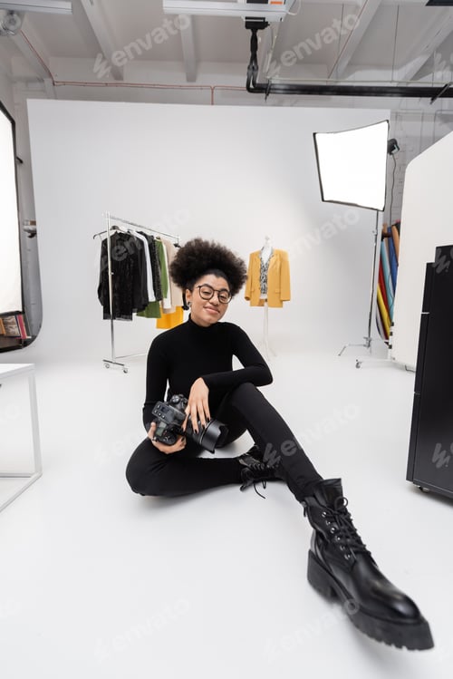 Preview: african american content producer in eyeglasses and black clothes smiling at camera while sitting in