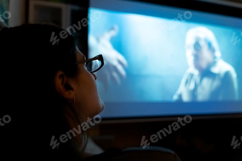 Preview: Young woman, girl with glasses watches tv, movie at night on huge screen at home on couch in room. I