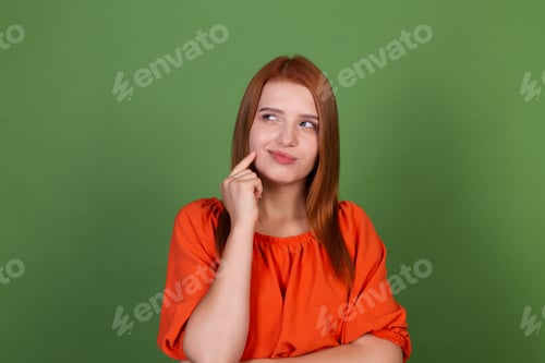 Preview: Young red hair woman in casual orange blouse on green background thoughtful thinking