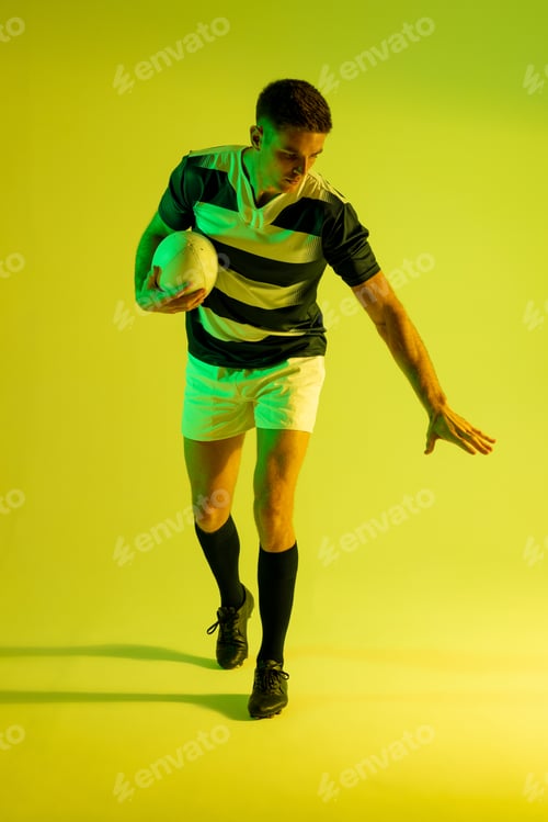 Preview: Caucasian male rugby player with rugby ball over yellow lighting