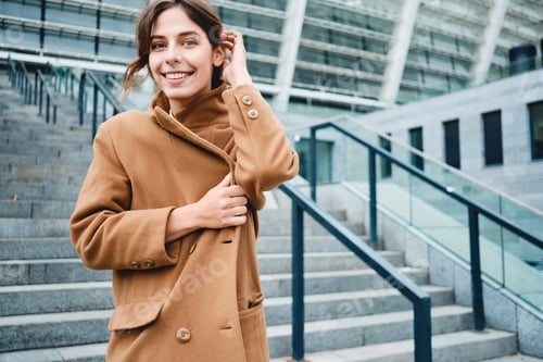 Preview: Young positive woman fastening up coat from cold happily looking in camera outdoor