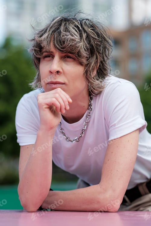 Preview: Thoughtful gender fluid person leaning on a pink surface outdoors