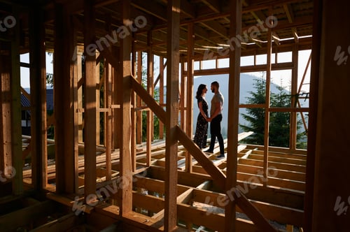 Preview: Man and woman inspecting their future wooden frame house nestled in the mountains near forest.