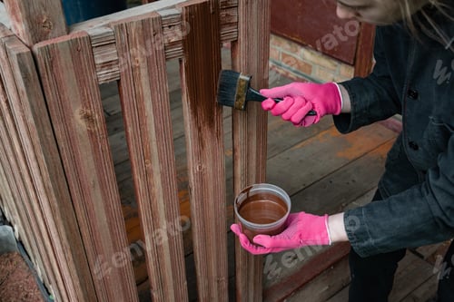 Preview: Person painting wooden fence with brush and container in hand on sunny day