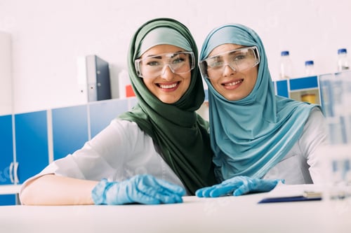 Preview: smiling female muslim chemists in protective goggles and hijab looking at camera in laboratory