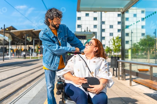 Preview: Young man assisting smiling woman in wheelchair at tram stop