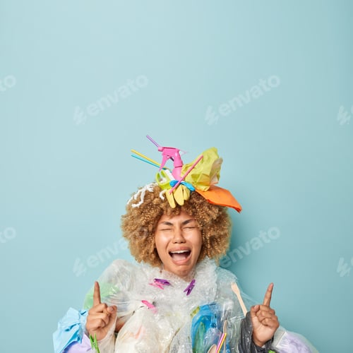 Preview: Upset crying female activist surrounded by plastic garbage points index fingers above feels worried