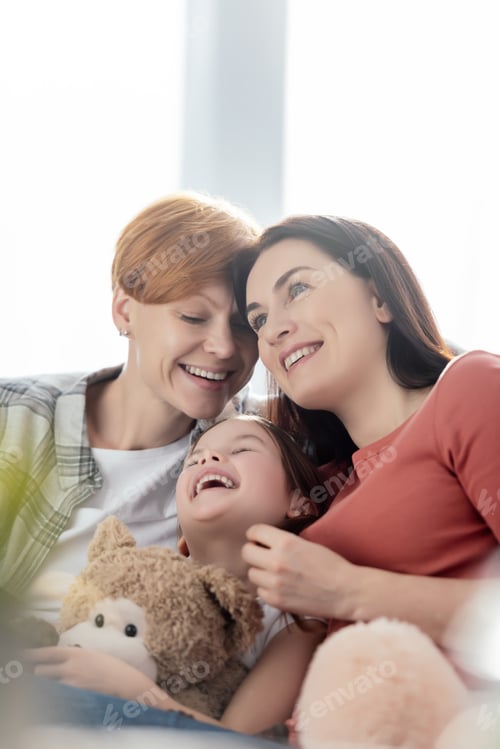 Preview: Selective focus of child with teddy bear laughing near smiling same sex parents at home