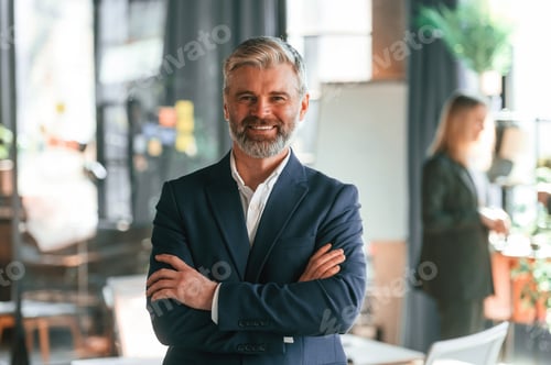 Preview: Senior man is standing with arms crossed. Business people are working together in the office