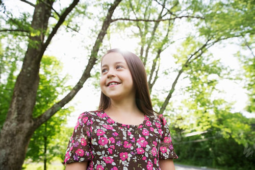 Preview: A young girl in a patterned summer dress, under the shade of trees in a farmhouse garden.