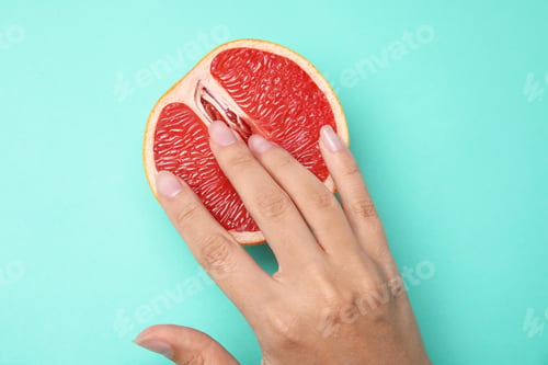 Vorschau: Junge Frau berührt eine halbe Grapefruit auf türkisfarbenem Hintergrund, Ansicht von oben. Sex-Konzept