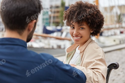 Preview: Content black teenage girl dressed in jacket, sits at bench together with handsome guy, pose togethe