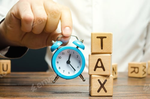 Preview: Businessman's hand holding a clock near the wooden blocks with the word Tax