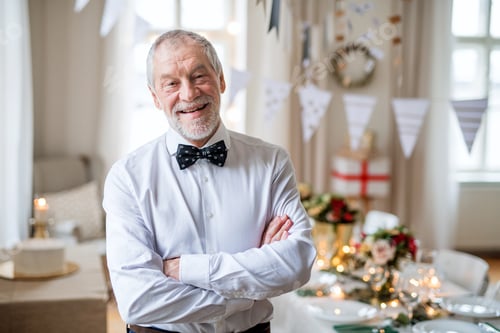 Preview: A portrait of a senior man standing indoors in a room set for a party, arms crossed.