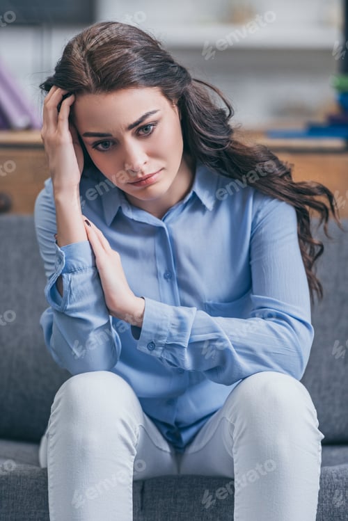 Preview: sad woman in blue blouse and white pants sitting on grey couch at home, grieving disorder concept