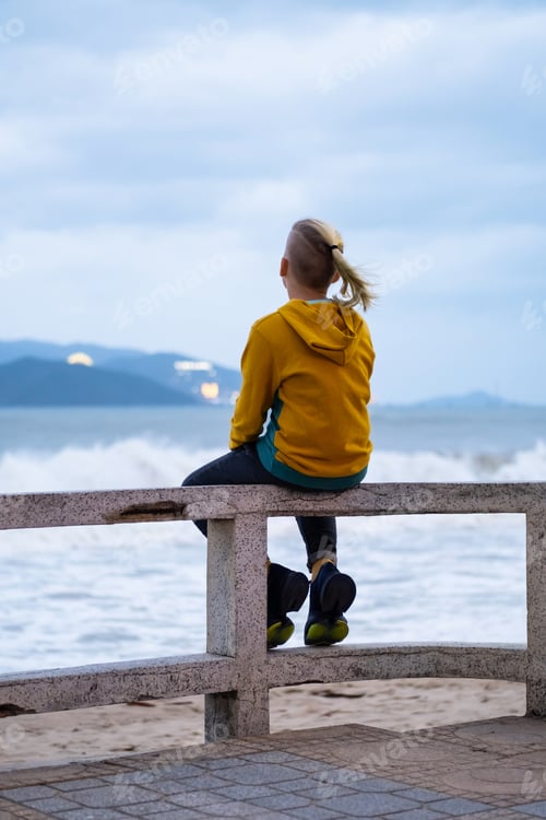 Preview: Lonely teenager looking to sea sitting on the waterfront. Rear view of teenager against seascape.
