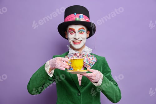 Preview: Positive crazy hatter wears bright colorful makeup enjoys drinking tea on party dressed in costume c