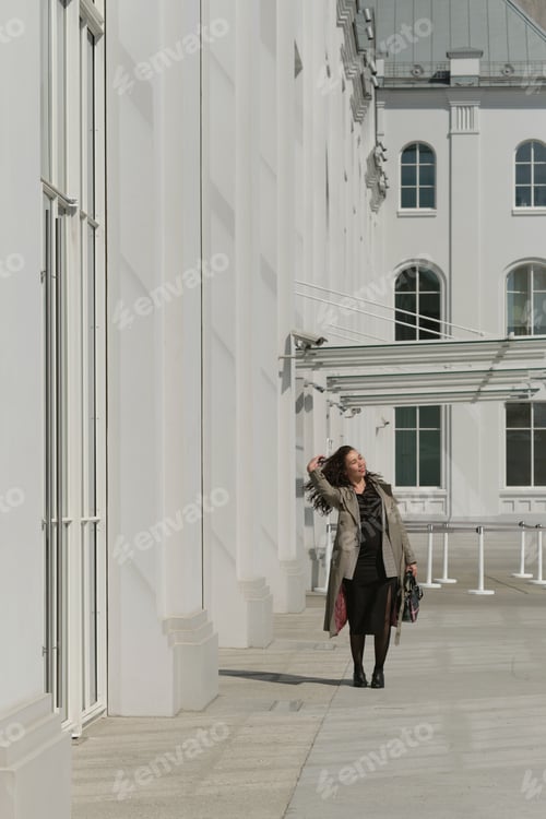Preview: Woman in a trench coat walking along a modern white building, enjoying the sunny day. Concept