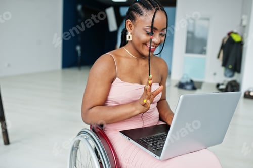 Preview: Young disabled African American woman in wheelchair at home working with laptop.