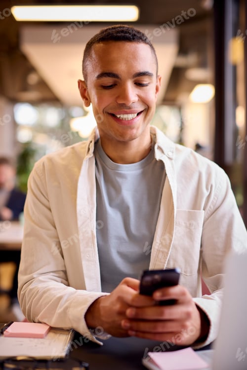 Preview: Young Businessman Working On Laptop At Desk In Office Checking Messages On Mobile Phone