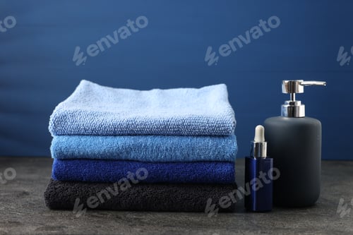 Preview: Stack of fresh towels and cosmetic products on grey table against blue background, closeup