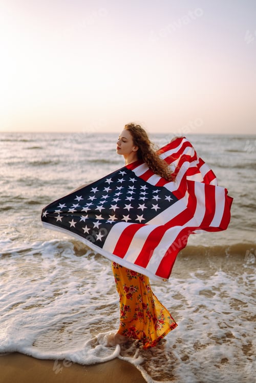 Preview: Young woman with american flag on the beach at sunset. 4th of July. Independence Day.
