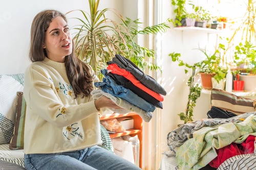 Preview: Caucasian young woman with long hair folding clean clothes sitting in the living room at home