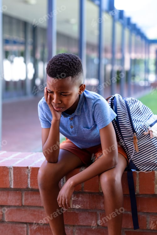 Preview: Vertical of sad african american schoolboy sitting on wall outside school, with copy space