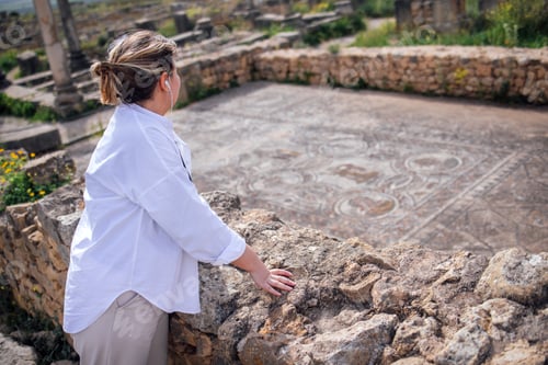 Preview: Woman in white shirt stands by ancient stone wall, gazing at historical ruins