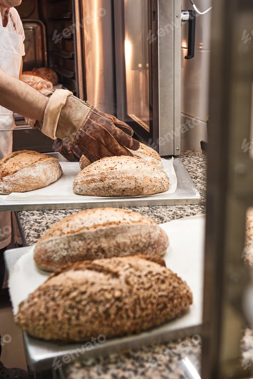 Preview: Freshly baked bread. Vertical photo of baker's hands in working gloves taking out hot bread from