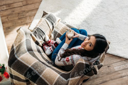 Preview: top view of young woman enjoying hot chocolate with marshmallow while sitting in cozy armchair