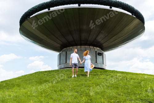 Preview: redhead couple holding hands and walking on grassy hill in front of modern building