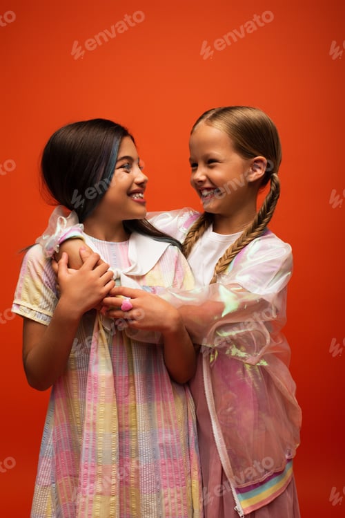 Preview: happy preteen girls in stylish clothes embracing and looking at each other isolated on orange