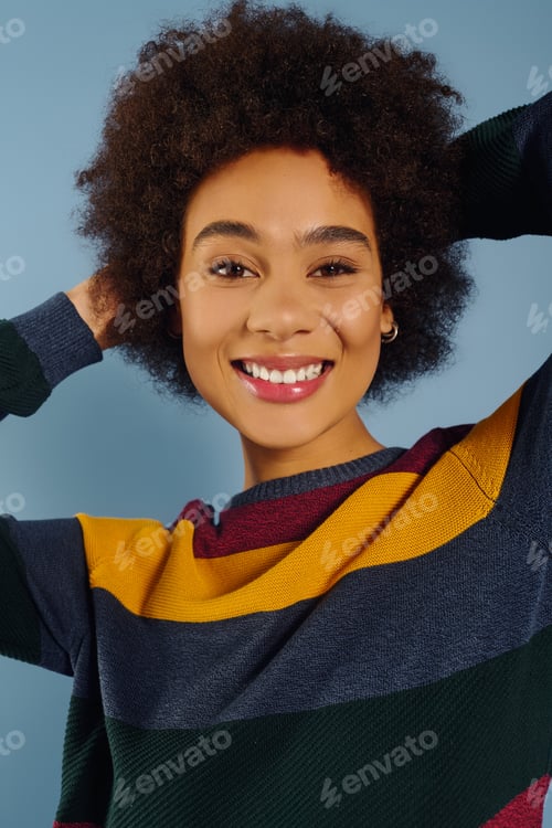 Preview: Joyful young woman with natural hair smiles brightly in a vibrant sweater against a blue backdrop