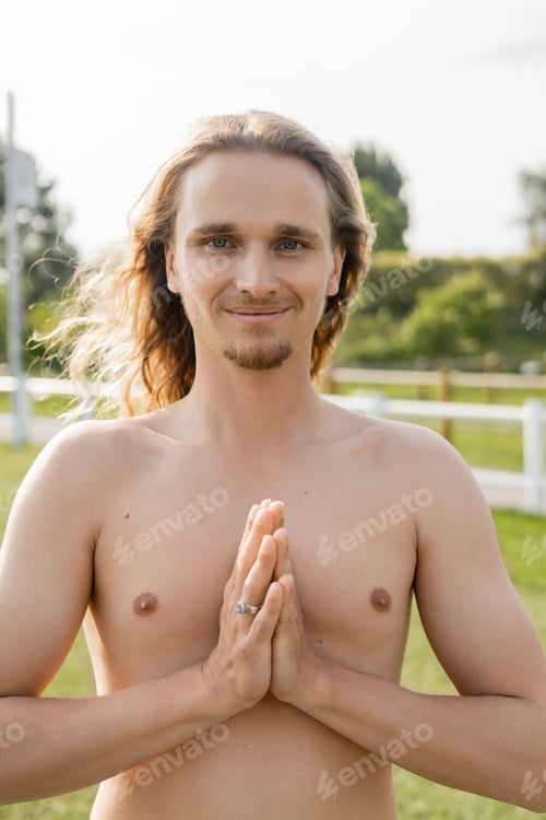 Preview: overjoyed shirtless man with long hair showing anjali mudra gesture and smiling at camera outdoors
