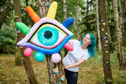 Preview: Smiling young adult woman with dyed turquoise hair in white T shirt near big eye art installation