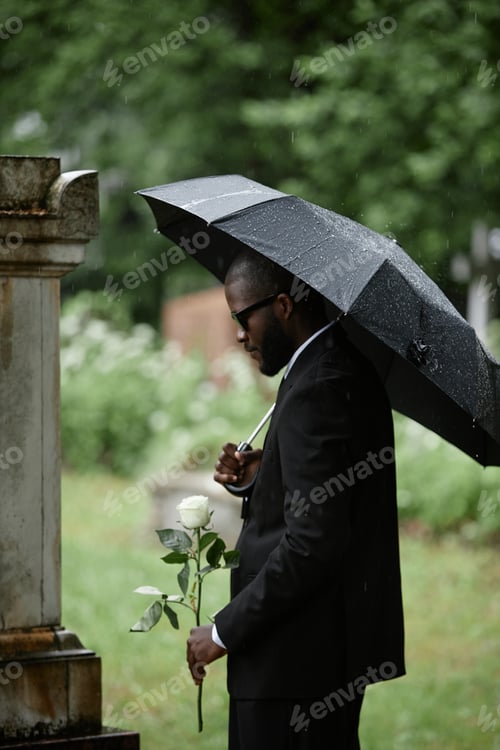 Preview: Black Man Grieving next to Grave Holding Rose under Umbrella at Graveyard