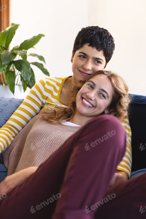 Preview: Happy biracial lesbian couple relaxing on couch together embracing and smiling at home