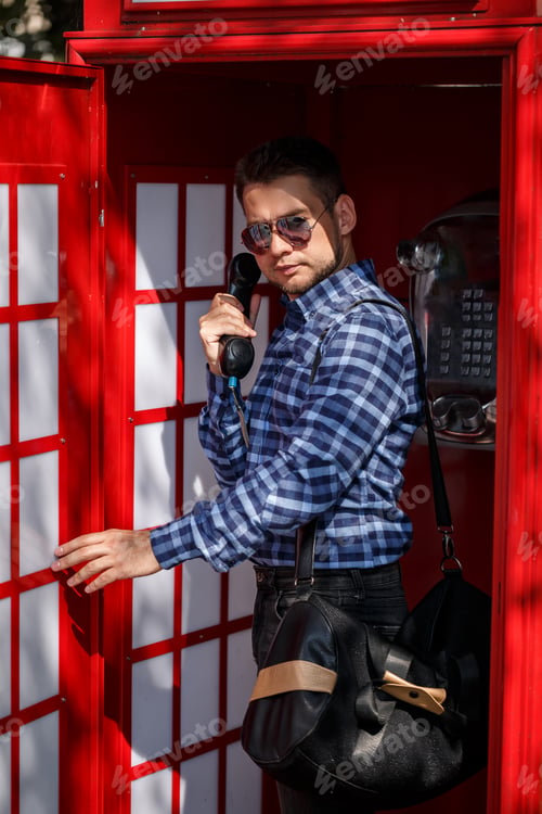 Preview: A handsome male businessman with a beard and a bag is standing in a red telephone booth and talking