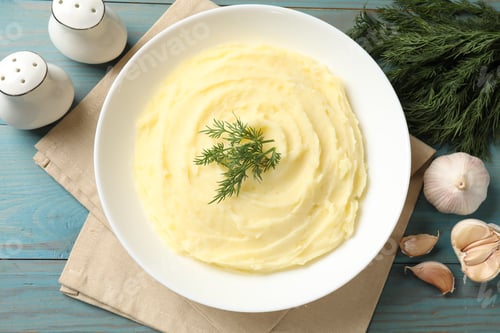 Preview: Tasty mashed potato with dill in bowl, garlic and spices on light blue wooden table, flat lay