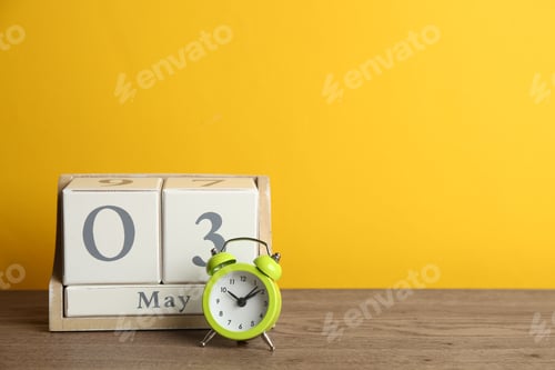 Preview: Wooden block calendar and alarm clock on table against yellow background. Space for text