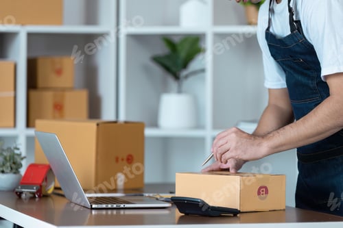 Preview: Small Business Entrepreneur Packing Orders in Home Office with Laptop and Cardboard Boxes for Online