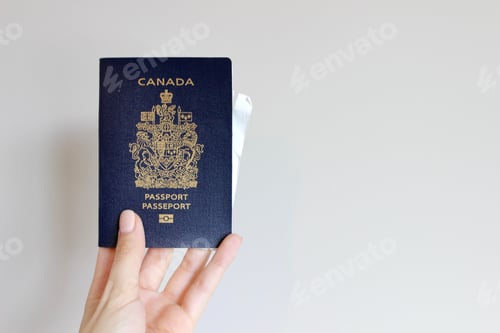 Preview: Woman’s hand holding a navy blue Canadian passport for traveling against a plain white wall